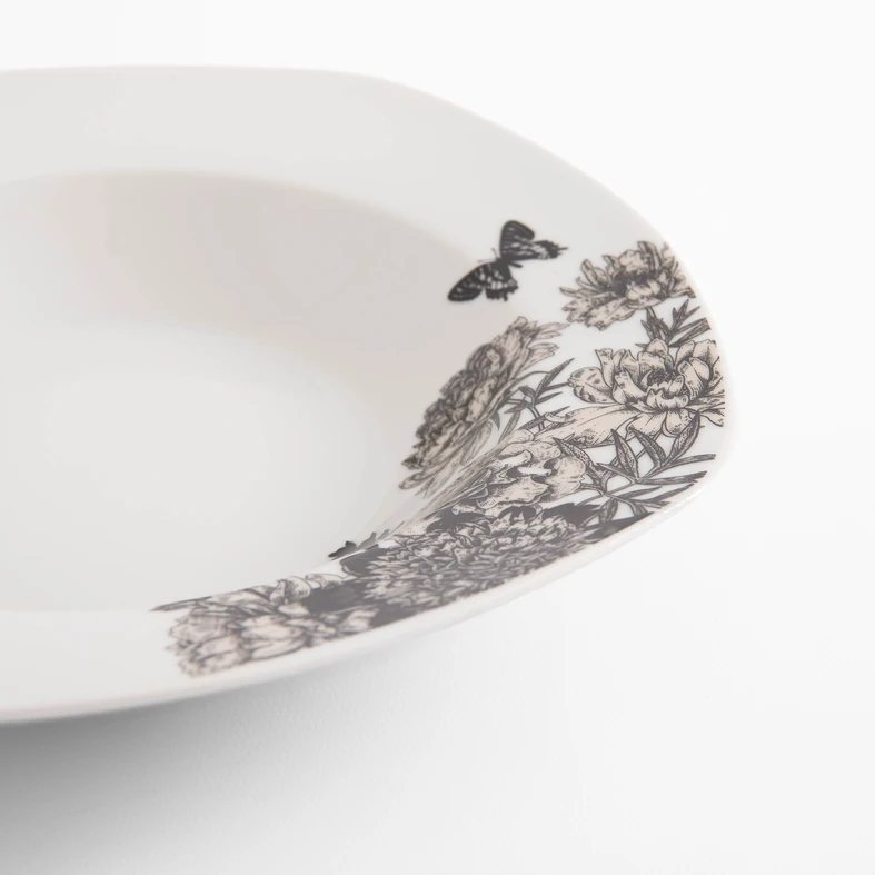 Detal, krawędź białego, porcelanowego talerza, widoczny delikatny kwiatowy wzór. Rysunek kwiatów piwonii i wirujących wokół nich motyli, uzupełniony kolorem jasnej sepii. 