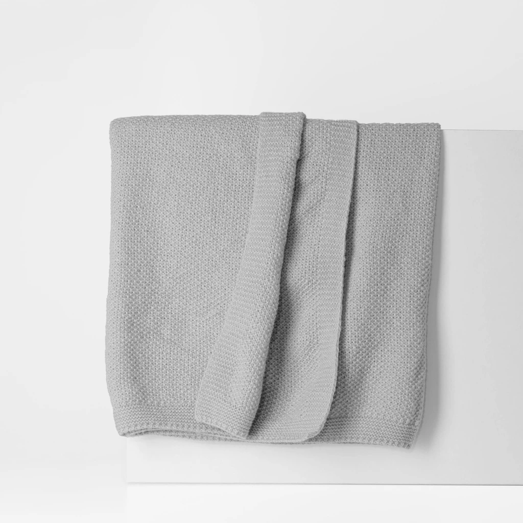 Szary koc o delikatnej, dzianinowej strukturze. O prostych, estetycznie wykończonych krawędziach.