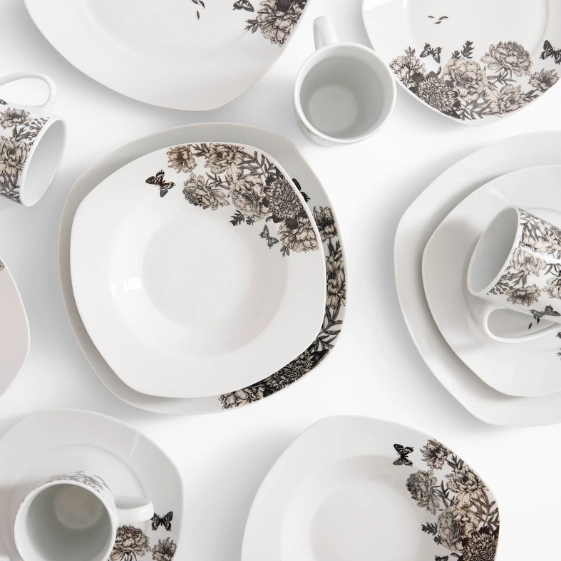 Rzut z góry, zestaw obiadowy z delikatnym kwiatowym wzorem na białej porcelanie. Widoczne talerze obiadowe, głębokie i deserowe oraz kubki. Naczynia mają lekko kwadratowy kształt.