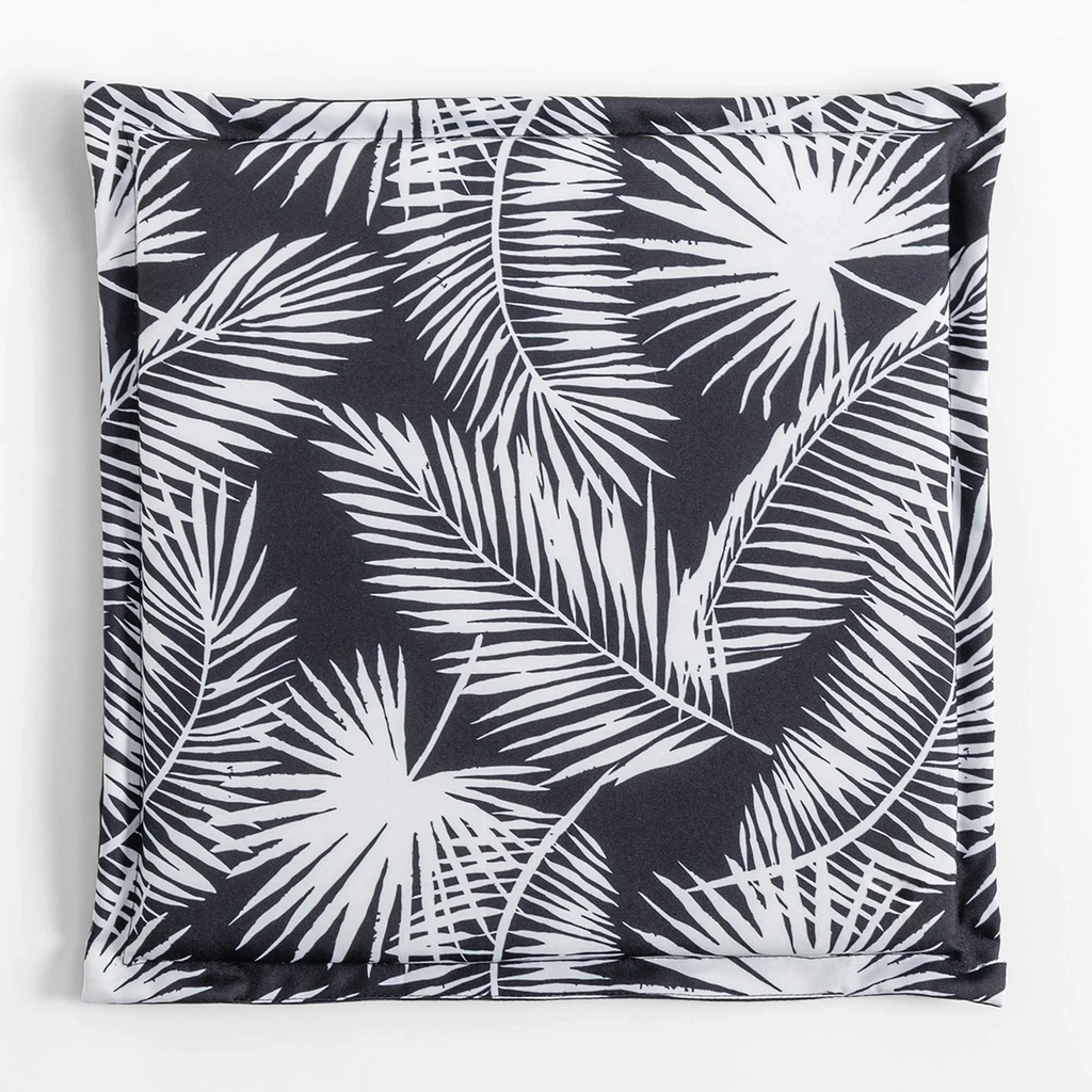 Kwadratowa, płaska poduszka ogrodowa, o wzorze białych liści palmowych na czarnym tle, o gładkim, czarnym tyle.