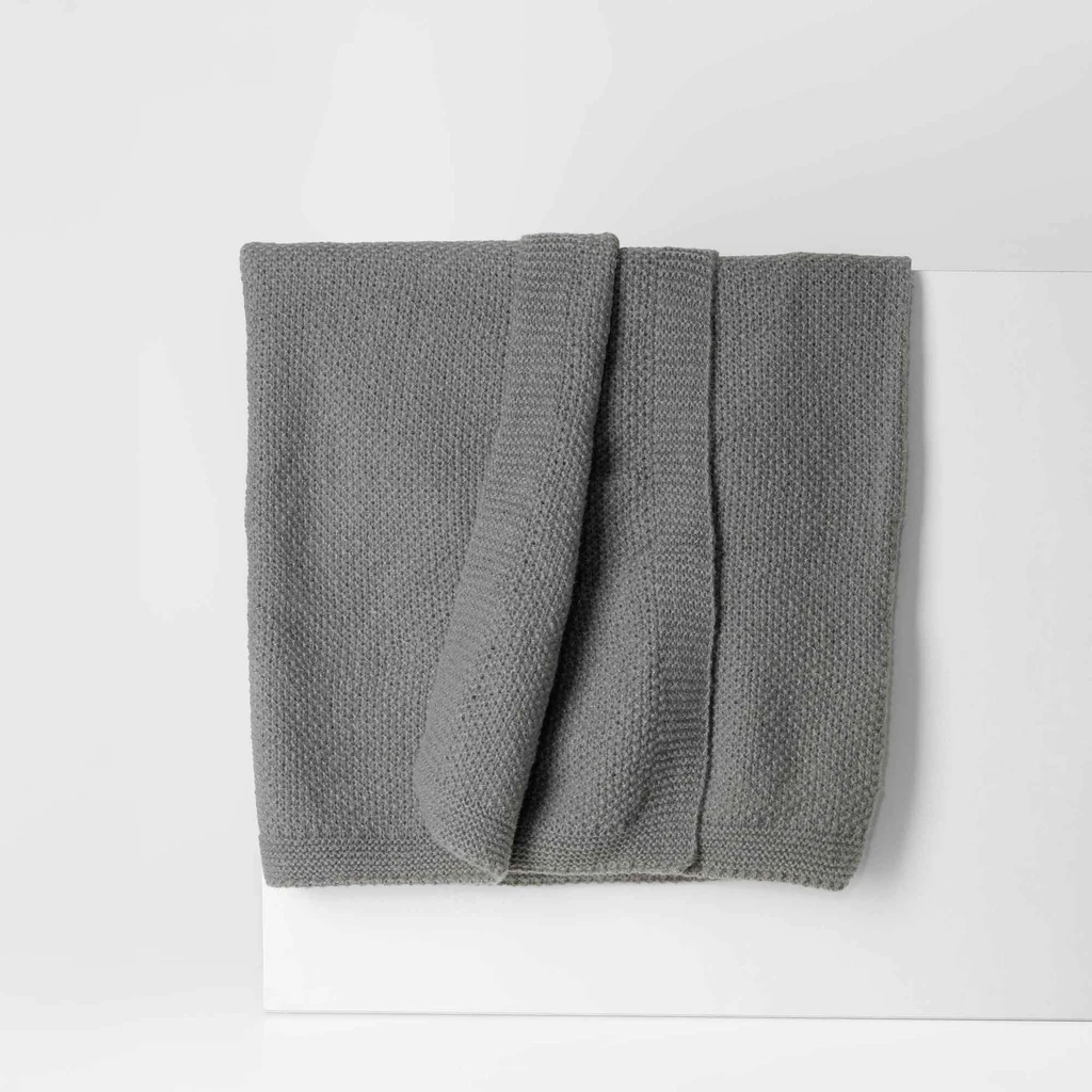 Ciemnoszary koc o delikatnej, dzianinowej strukturze. O prostych, estetycznie wykończonych krawędziach.