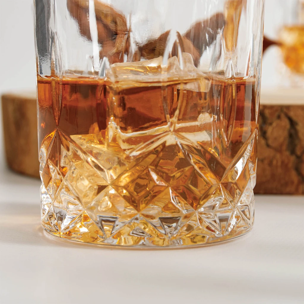 Zbliżenie na podstawę szklanki do połowy wypełnionej whisky. Bursztynowy napój podkreśla wzór kryształowego szlifu. 