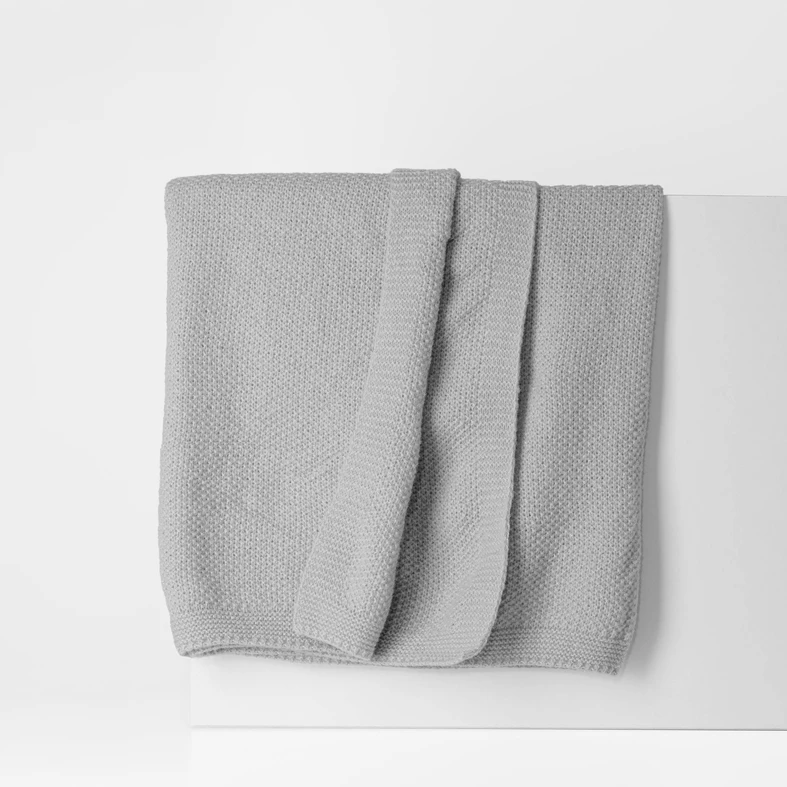 Szary koc o delikatnej, dzianinowej strukturze. O prostych, estetycznie wykończonych krawędziach.