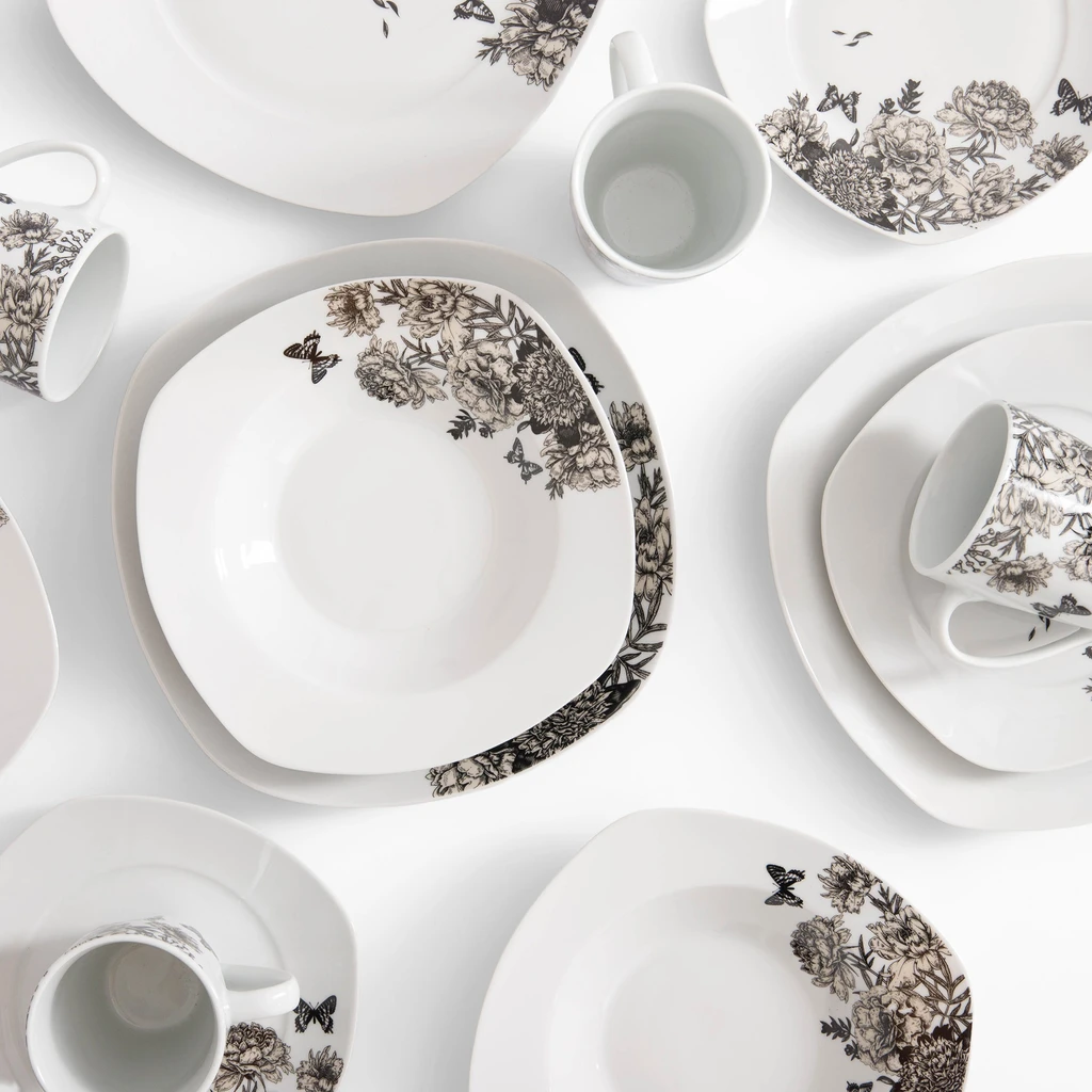 Rzut z góry, zestaw obiadowy z delikatnym kwiatowym wzorem na białej porcelanie. Widoczne talerze obiadowe, głębokie i deserowe oraz kubki. Naczynia mają lekko kwadratowy kształt.