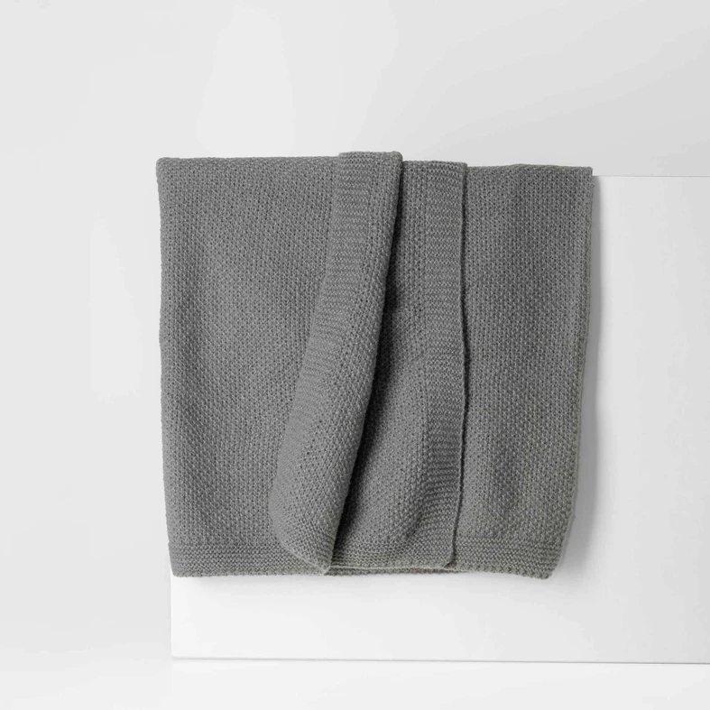 Ciemnoszary koc o delikatnej, dzianinowej strukturze. O prostych, estetycznie wykończonych krawędziach.