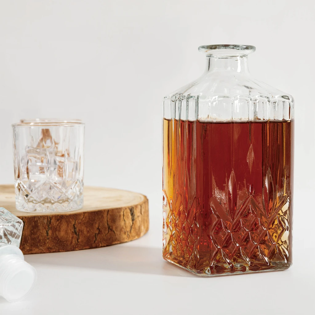 Szklana karafka wypełniona płynem w kolorze bursztynu. Obok niej szklanka z kostkami lodu, umieszczona na drewnianym krążku.
