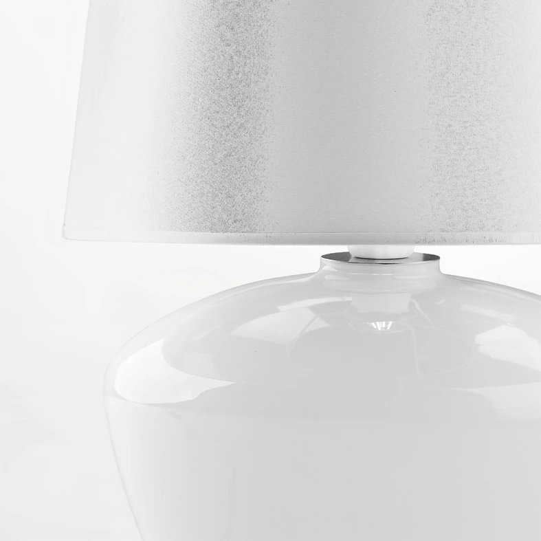 Abażur ma klasyczny kształt trapezu, opinająca go tkanina jest biała.