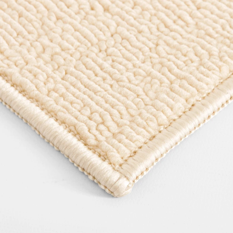 Zbliżenie na gęstą, pętelkową powierzchnię beżowego dywanika łazienkowego ze starannie obszytym brzegiem.